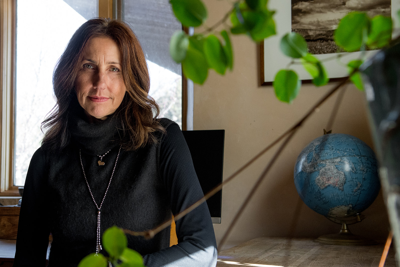 Linda Seebantz in a black turtleneck by a sunlit window in her studio, a globe and green leaves beside her